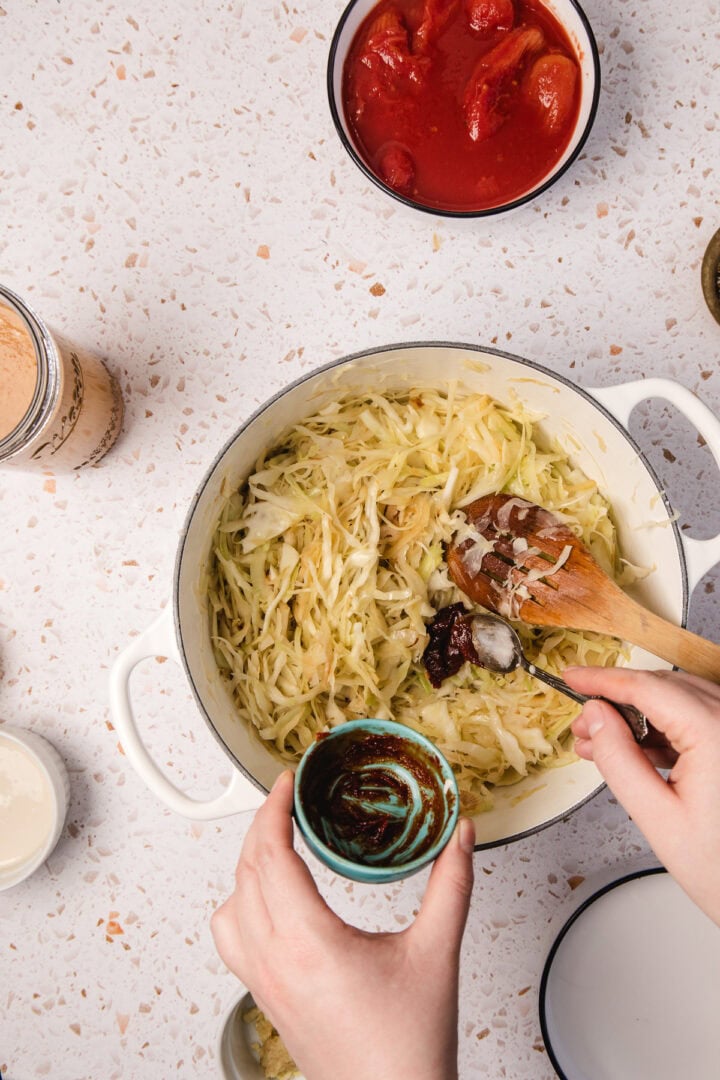 Hands adding sauce to shredded cabbage in a white pot, surrounded by bowls of ingredients&mdash;preparing flavorful Gochujang Tahini Cabbage Soup on the countertop.