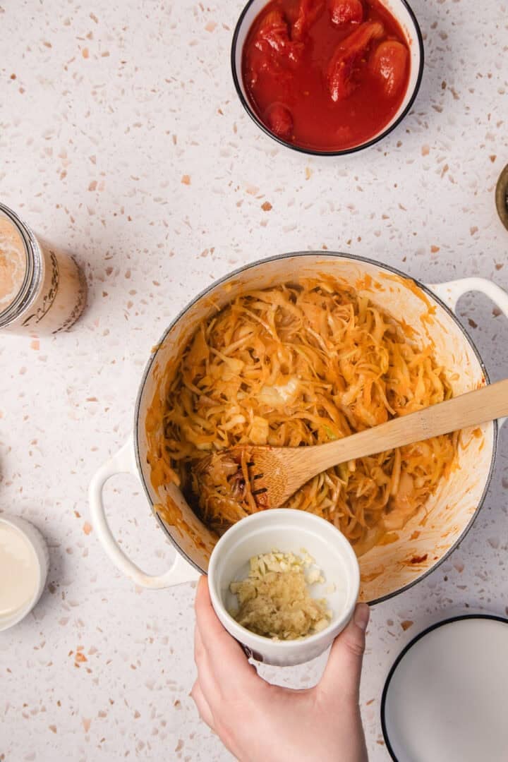 A hand holds a bowl of minced garlic above a pot of Gochujang Tahini Cabbage Soup, filled with shredded cabbage in sauce and stirred with a wooden spoon.