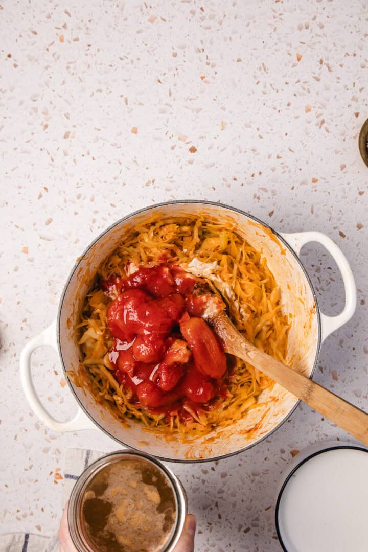 A pot of Gochujang Tahini Cabbage Soup is being stirred, filled with shredded vegetables and whole tomatoes, as a hand hovers above holding a can on the countertop.