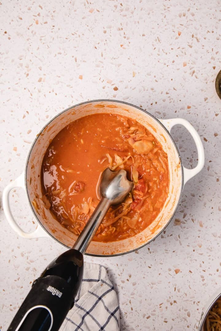 An immersion blender blending Gochujang Tahini Cabbage Soup in a white pot on a speckled countertop.