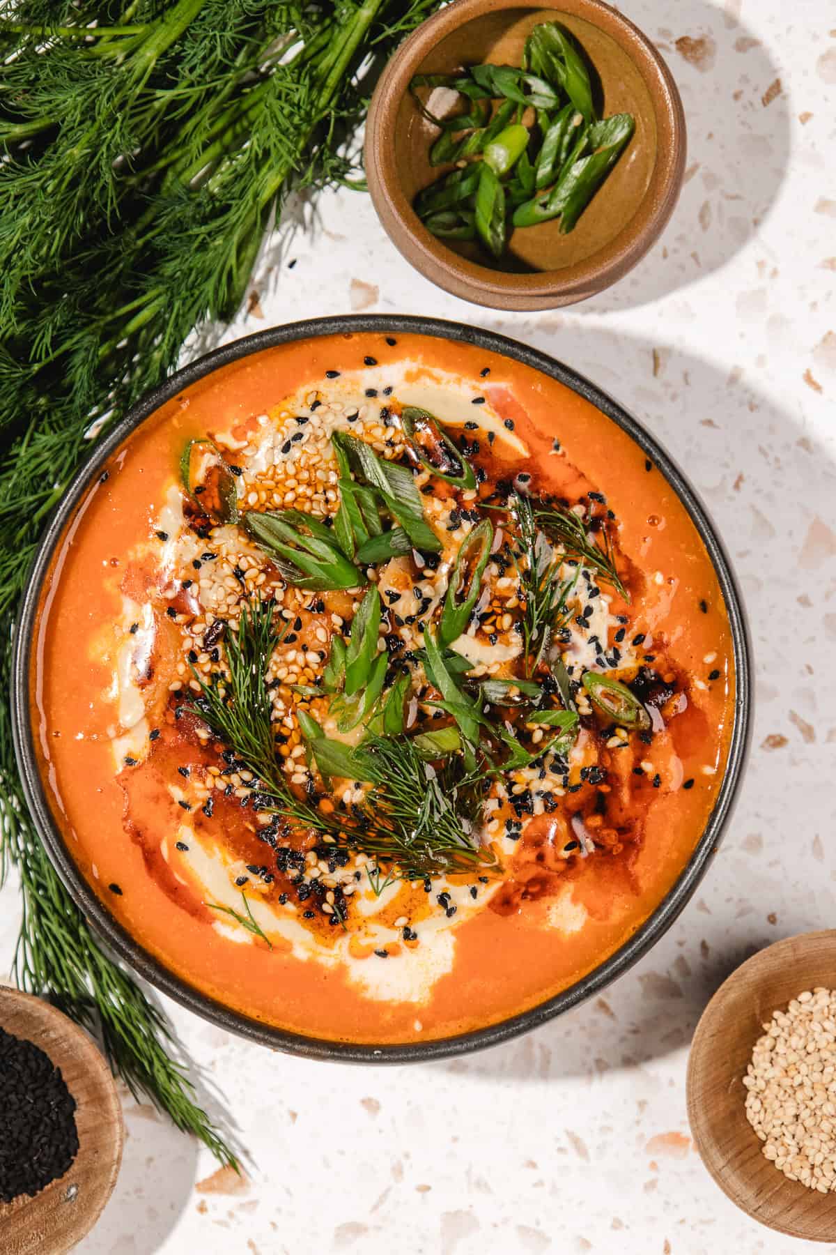 A bowl of creamy tomato soup, inspired by Gochujang Tahini Cabbage Soup, topped with herbs, seeds, green onions, and surrounded by fresh dill and seeds.