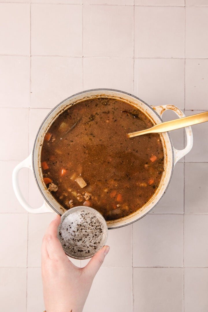 A hand adds seasoning to a pot of soup with vegetables on a tiled surface, with a wooden spoon in the pot.