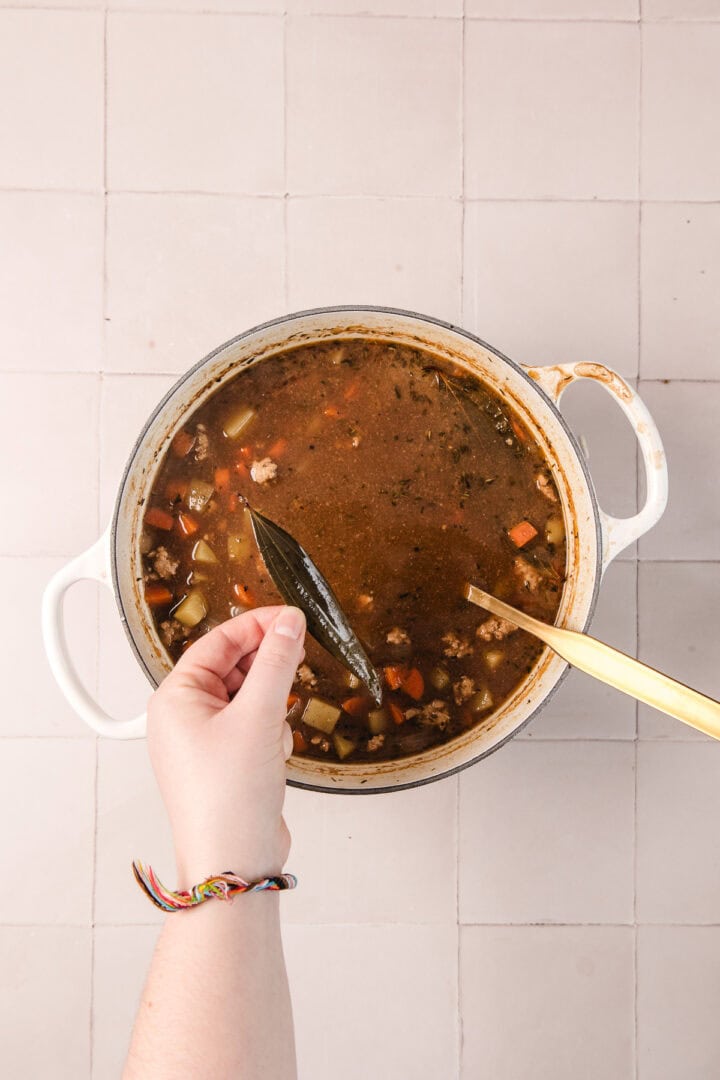 Hand dropping a bay leaf into a pot of vegetable soup on a tiled surface.