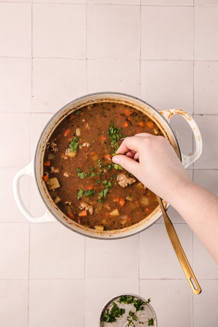 A hand sprinkles fresh herbs into a pot of vegetable soup on a tiled countertop.