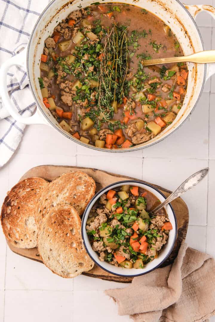 A pot of vegetable stew with herbs, a bowl of stew, and three slices of toasted bread on a wooden board.