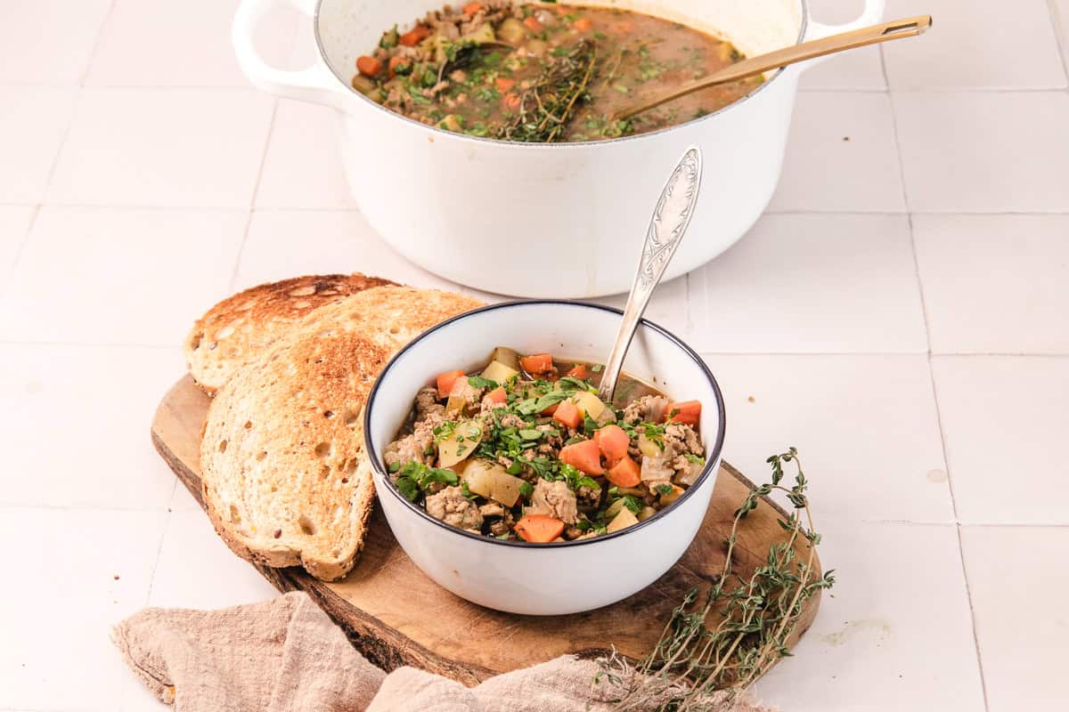A bowl of vegetable soup with bread slices on a wooden board, and a pot of soup in the background.