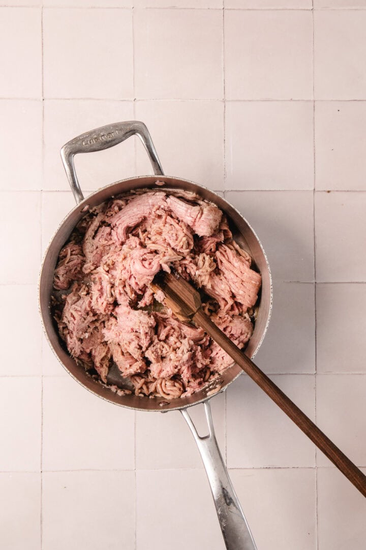 Shredded meat in a pot with a wooden spoon, on a light tiled surface.