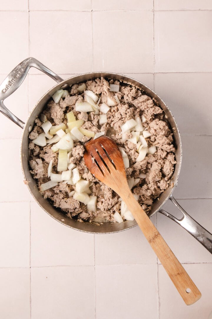 A skillet with ground meat and chopped onions being stirred with a wooden spoon on a tiled surface.