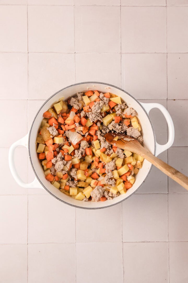A white pot with diced potatoes, carrots, and ground meat, stirred with a wooden spoon on a tiled surface.