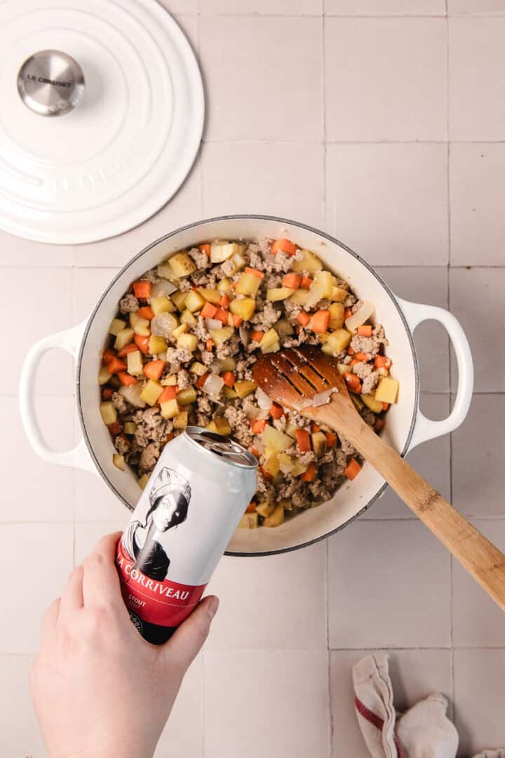 A hand pours beer from a can into a pot of diced vegetables and ground meat on a tiled countertop.