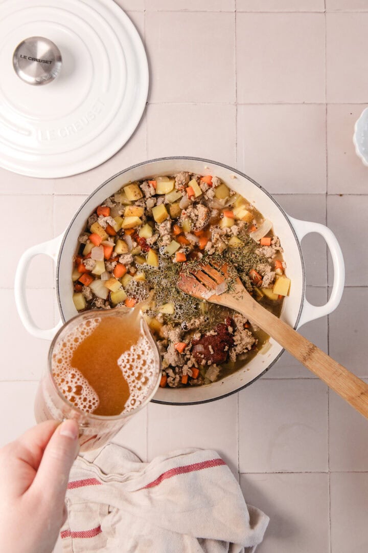A hand pours broth into a pot of diced vegetables and meat on a tiled surface, with a wooden spatula inside.