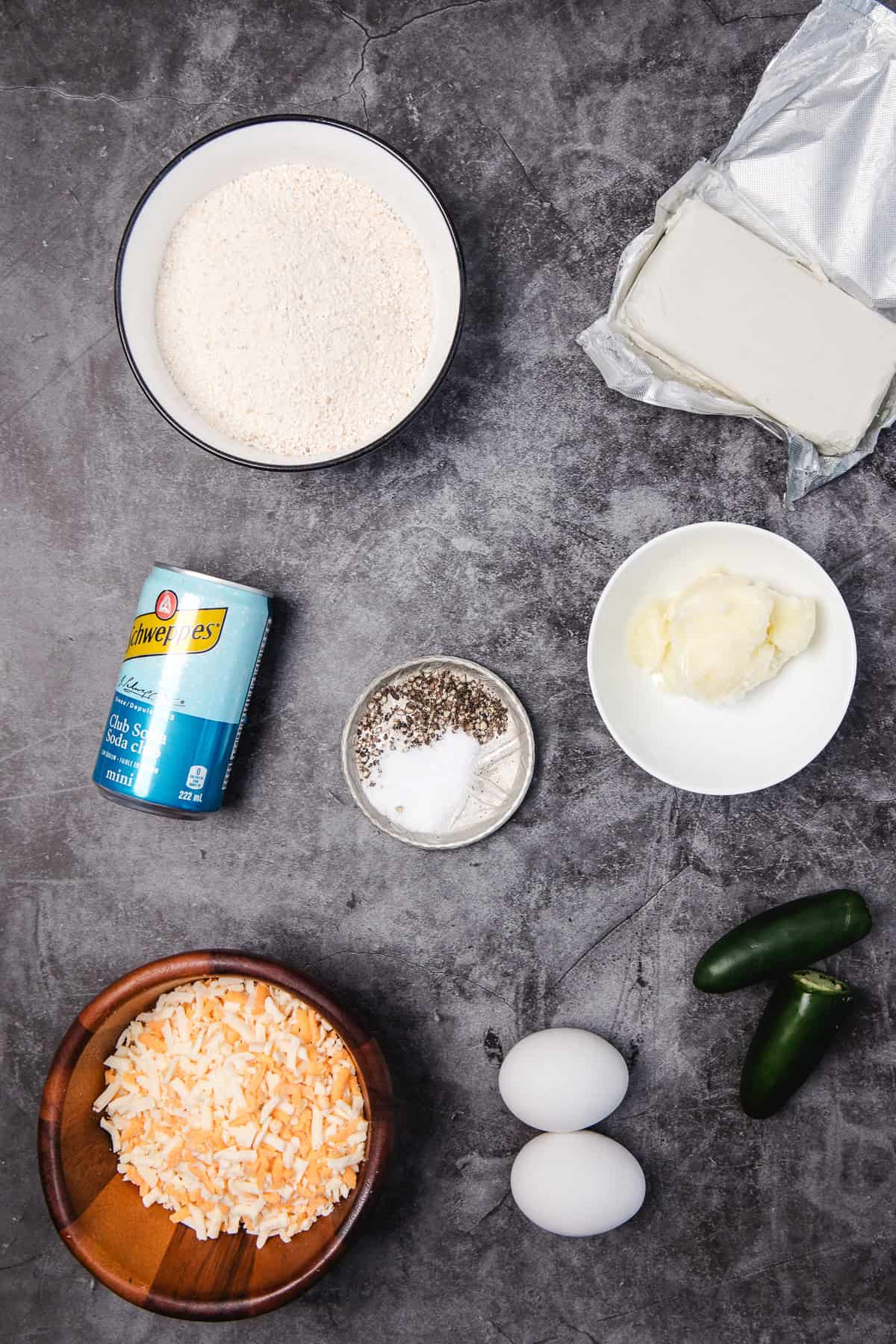 Ingredients on a gray surface: eggs, shredded cheese, jalapeños, cream cheese, butter, flour, and a can of baking powder.