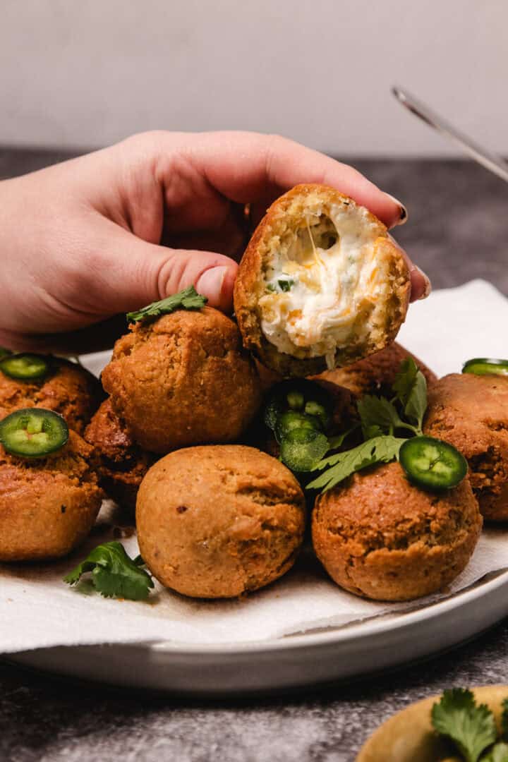 A hand holding a fried ball with creamy filling, next to more on a plate, garnished with jalapeño and cilantro.