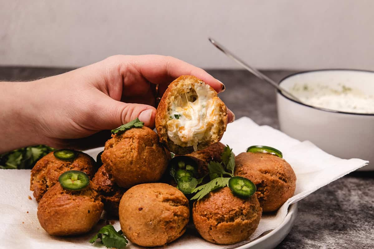 A hand holds a jalapeño popper with creamy filling, surrounded by more poppers garnished with cilantro and jalapeño.
