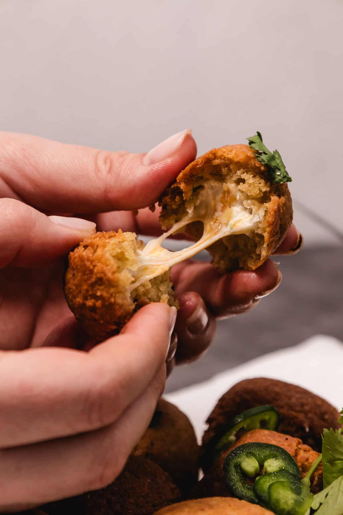 A hand pulls apart a fried ball revealing melted cheese inside; jalapeños and herbs are visible on others.