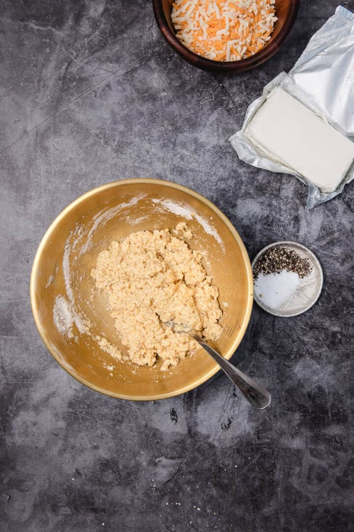 A mixing bowl with dough, a spoon, shredded cheese, cream cheese, salt, and pepper on a dark surface.
