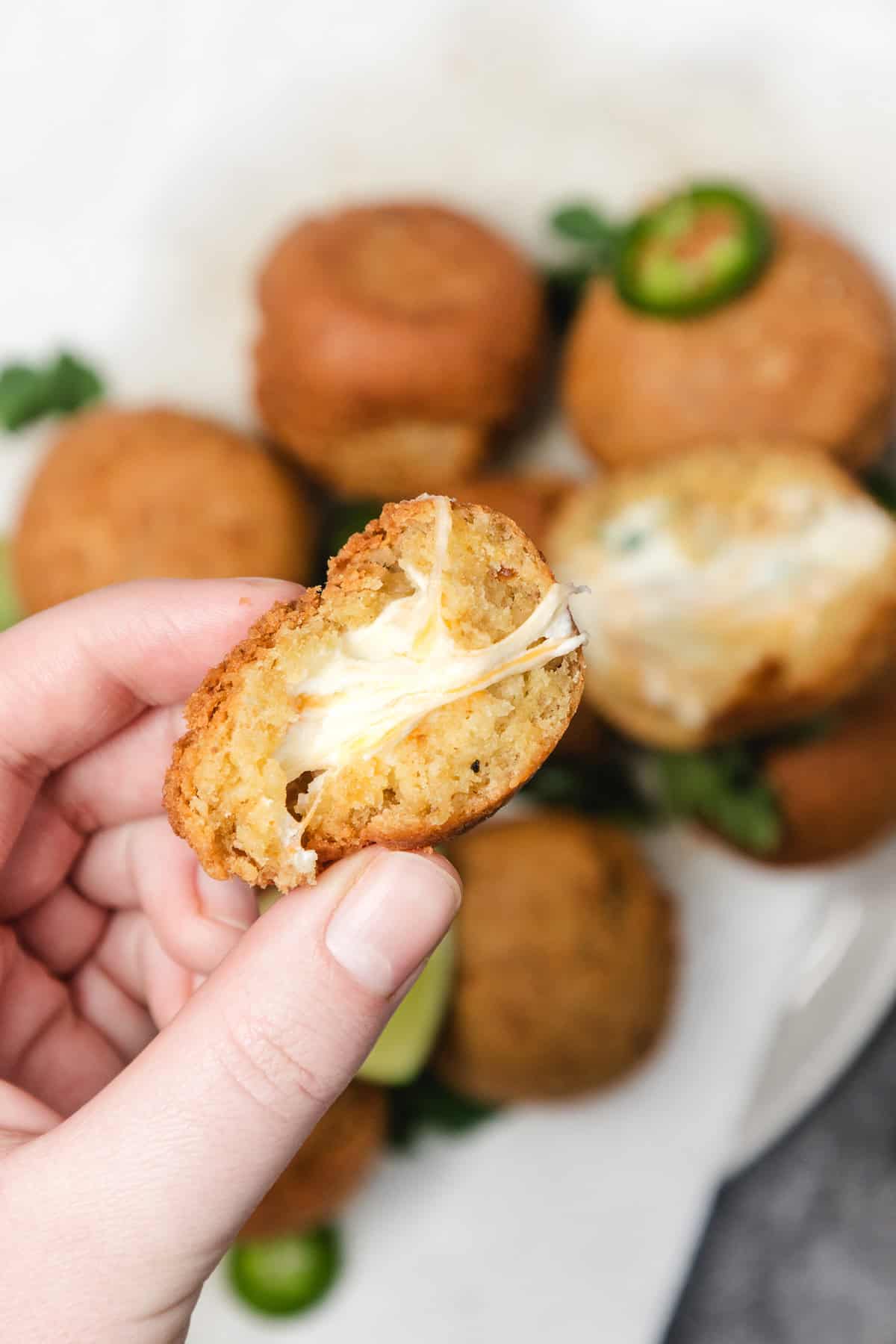 A hand holding a cheesy, half-eaten fried ball with jalapeños and more fried balls in the background.