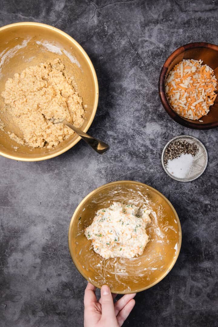 Three bowls with biscuit dough, shredded cheese, and seasonings on a dark countertop, hand holding one bowl.