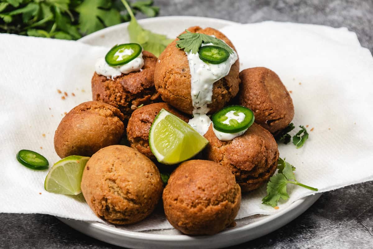 A plate of falafel balls with lime wedges, jalapeño slices, and white sauce, garnished with cilantro.