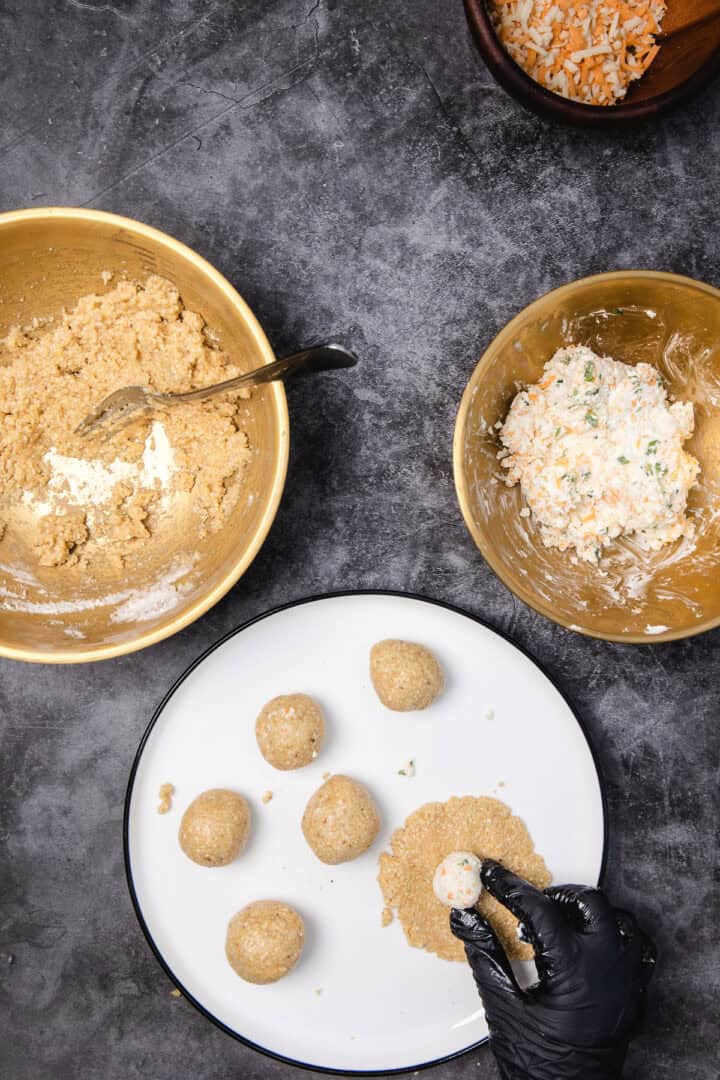 A gloved hand fills dough balls with a creamy mixture on a dark countertop, surrounded by mixing bowls.