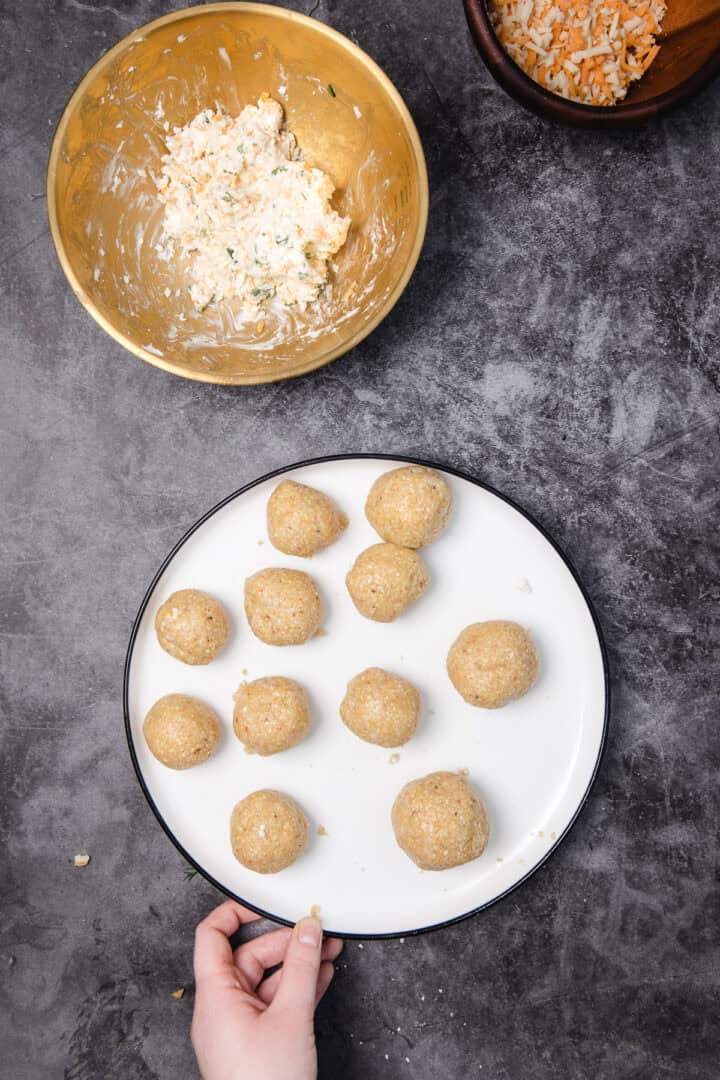 A hand holds a plate of round, breaded balls beside a bowl of creamy filling on a gray countertop.
