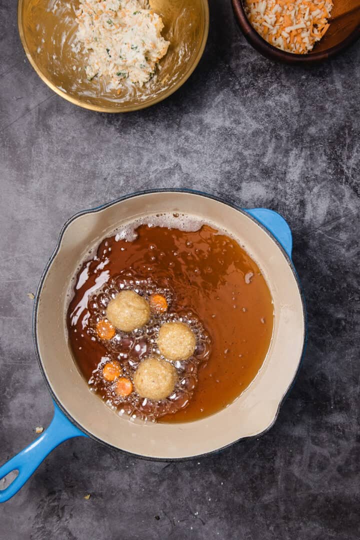 Four balls of dough are frying in hot oil in a blue pot, with bowls of ingredients nearby on a gray surface.
