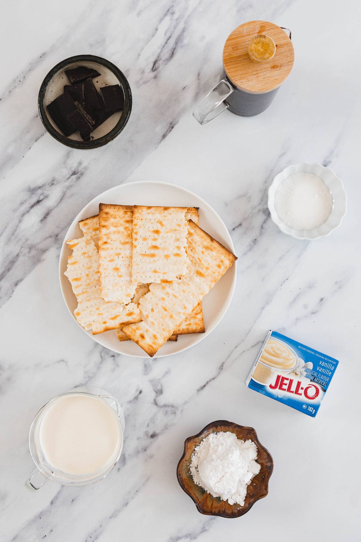 Top view of matzo crackers, chocolate, sugar, instant pudding, cream, and cornstarch on a marble surface.