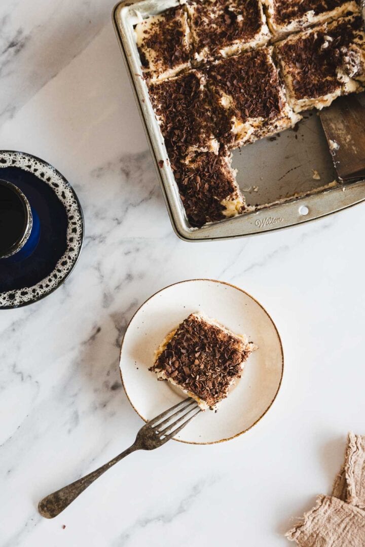 A slice of chocolate-topped dessert on a plate with a fork, next to a pan of similar dessert squares.
