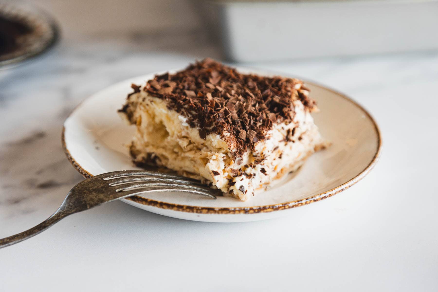 A slice of tiramisu topped with chocolate shavings on a plate with a fork beside it.