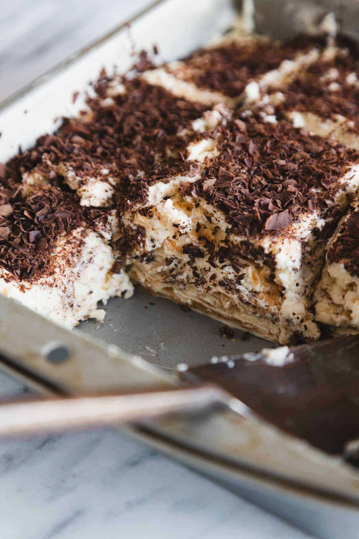 A close-up of tiramisu in a pan, topped with shaved chocolate and sliced to serve.