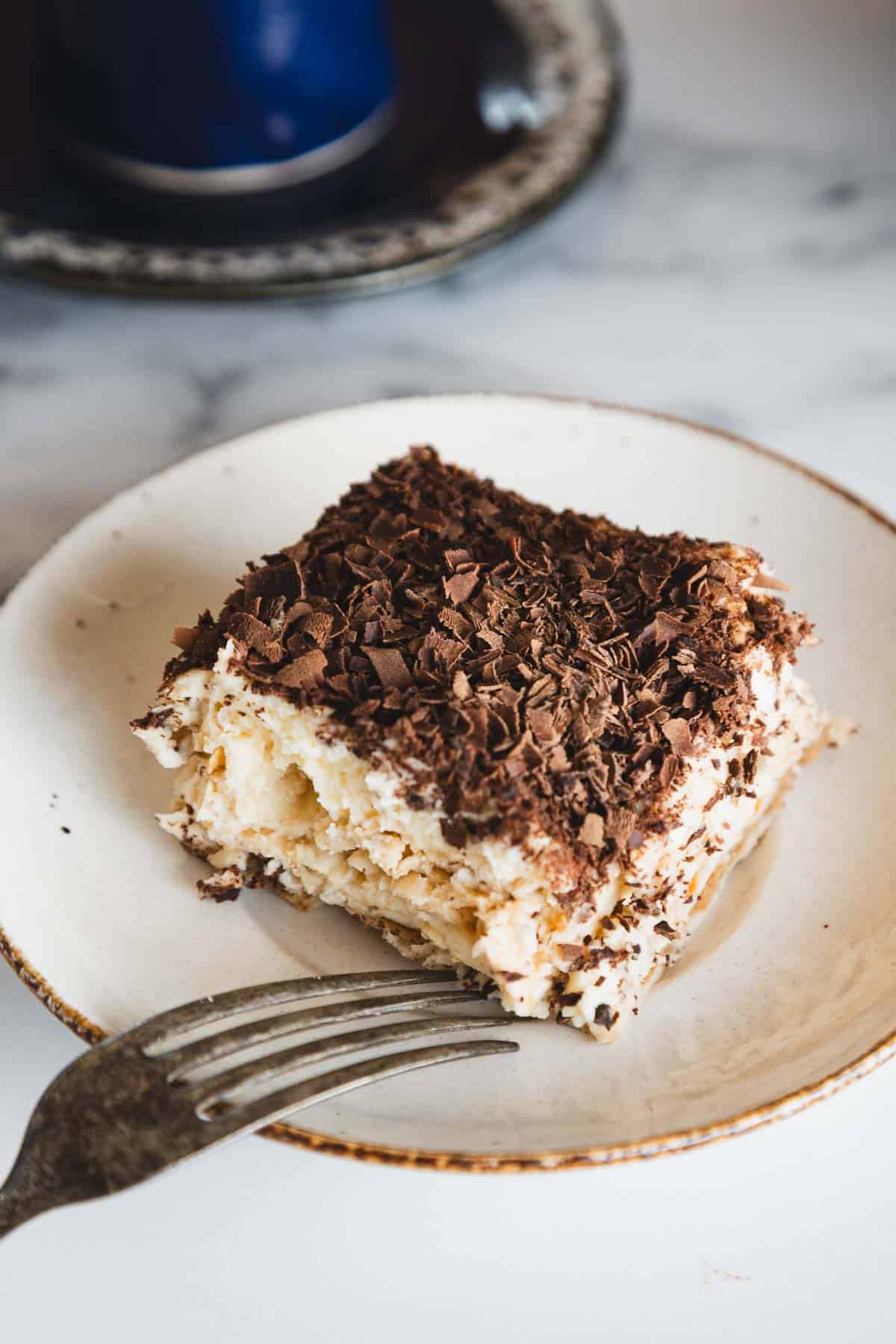 A slice of tiramisu topped with chocolate shavings on a plate, with a fork beside it.