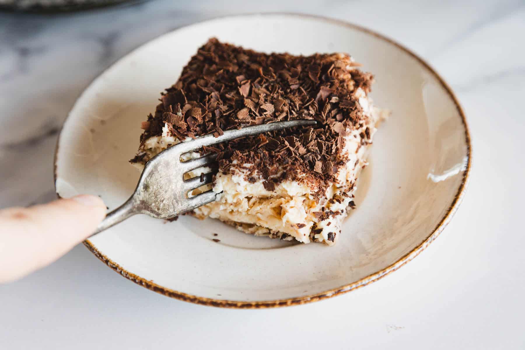 A fork cutting into a slice of creamy tiramisu topped with chocolate shavings on a plate.