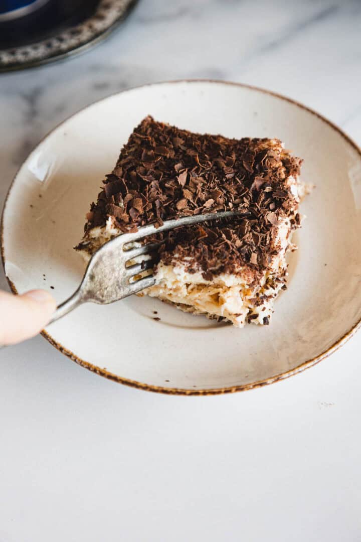 A fork cutting into a slice of tiramisu topped with chocolate shavings on a white plate.