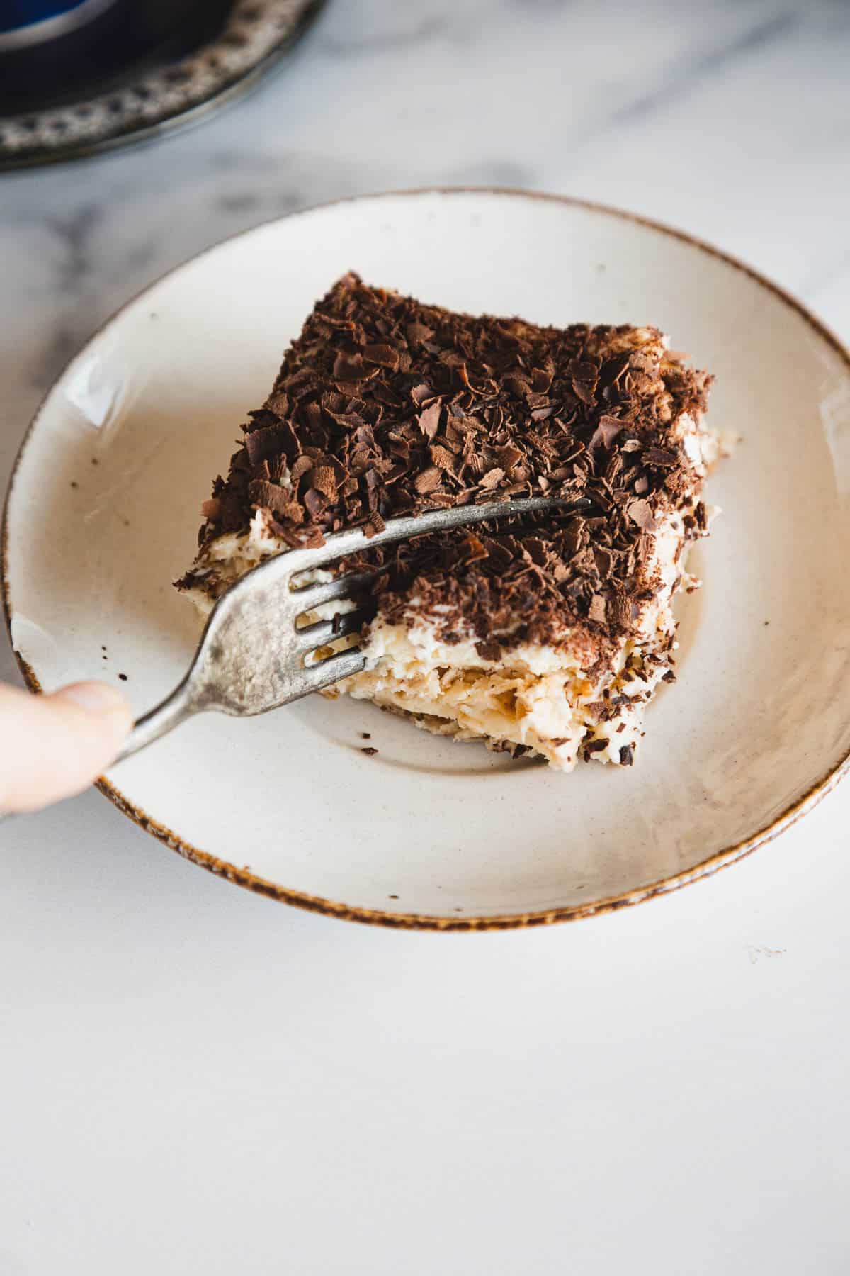A fork cutting into a slice of tiramisu topped with chocolate shavings on a white plate.