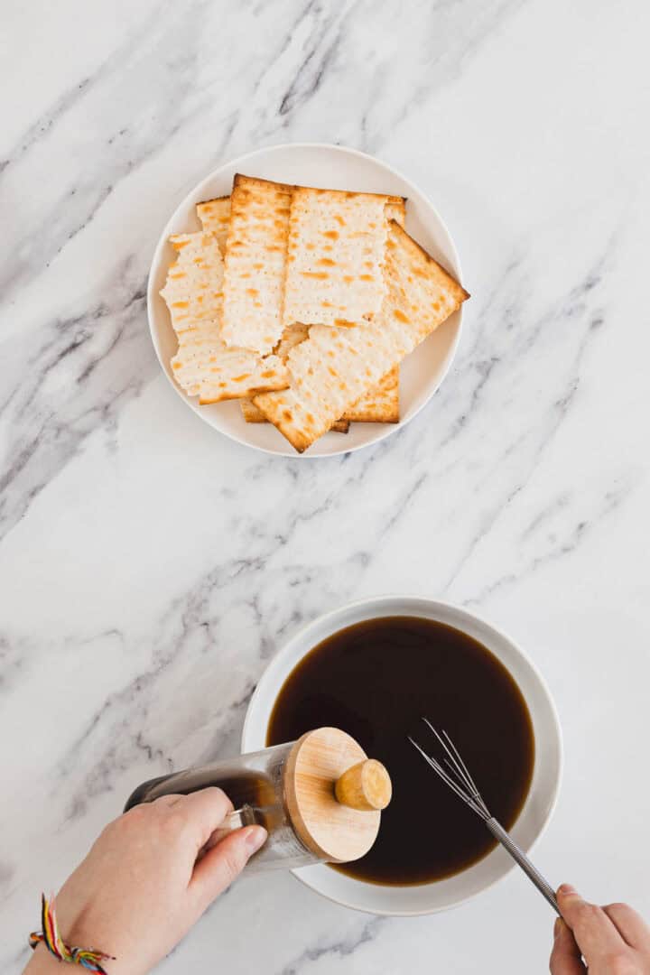 Plate of matzo crackers above a bowl of dark liquid, with hands holding a French press and a whisk.