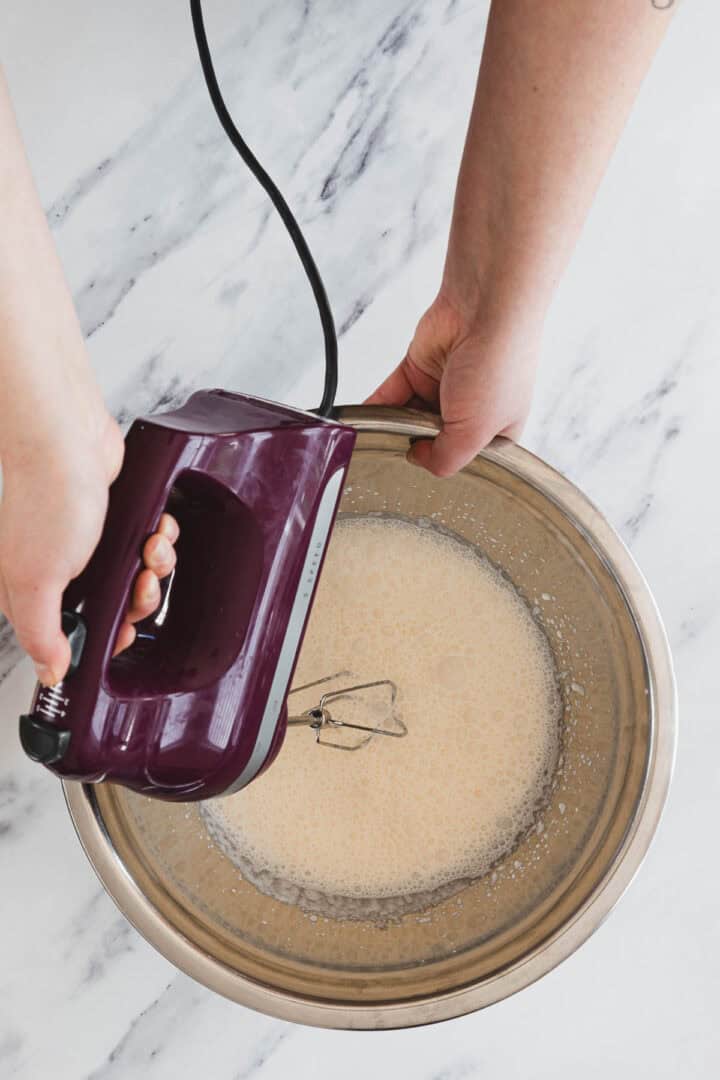 A person uses a hand mixer to blend ingredients in a metal bowl on a marble countertop.
