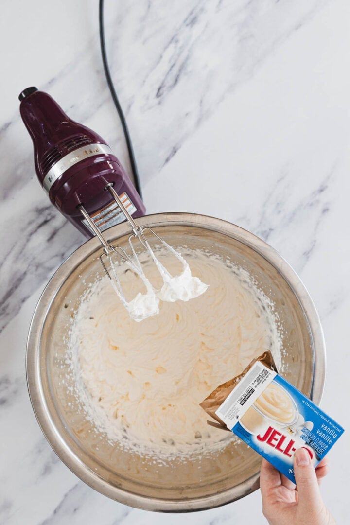 Hand holding a box of vanilla pudding over a mixing bowl with a hand mixer and whipped mixture inside.