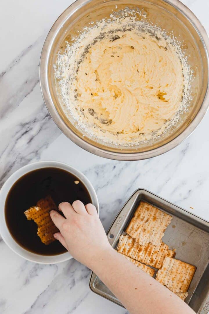 Hand dipping cracker into bowl of coffee, with whipped mixture and more crackers on a tray nearby.