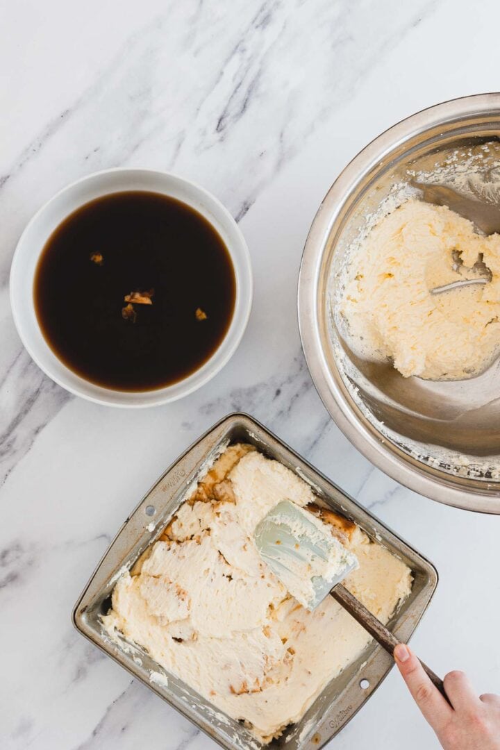 A hand spreads cream over dessert in a pan, next to a bowl of coffee and a bowl of cream mixture on a marble surface.
