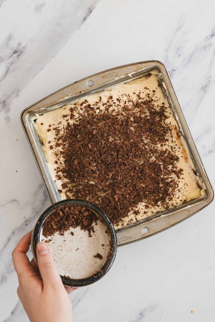 A hand sprinkles chocolate shavings onto a dessert in a square baking pan on a marble surface.