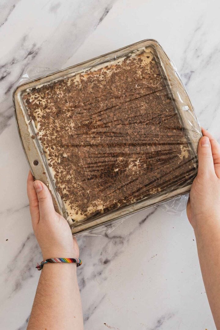Hands holding a square dish of tiramisu covered with plastic wrap on a marble countertop.