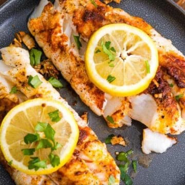 Two cooked fish fillets topped with lemon slices and herbs on a dark plate.