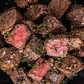 Steak bites topped with herbs and coarse salt on a black plate, some pieces are medium-rare.