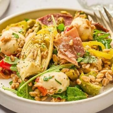 A plate of fresh salad with mozzarella, greens, prosciutto, grains, and sliced peppers, served with a fork.