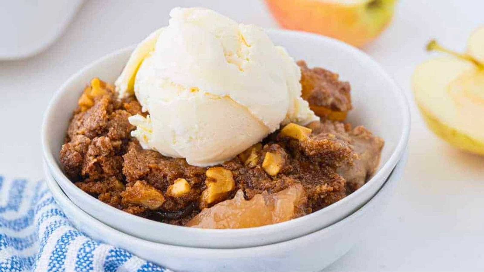 A bowl of apple crisp topped with vanilla ice cream, with apple slices in the background.