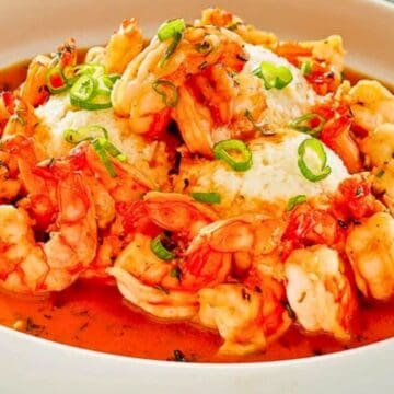 Shrimp in a red sauce, served with white dumplings and garnished with sliced green onions in a white bowl.