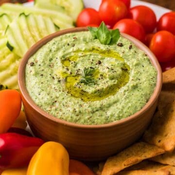 A bowl of green dip with olive oil, surrounded by crackers, cherry tomatoes, cucumbers, and mini peppers—perfect for serving as one of your favorite party dips.
