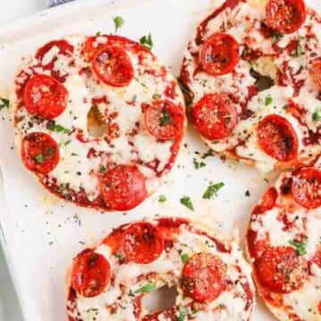 Four pepperoni pizza bagels topped with melted cheese and herbs on a white tray, next to a bowl of grated cheese.