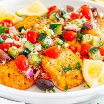 Grilled salmon fillets topped with a fresh cucumber, tomato, and olive salad, garnished with lemon wedges.