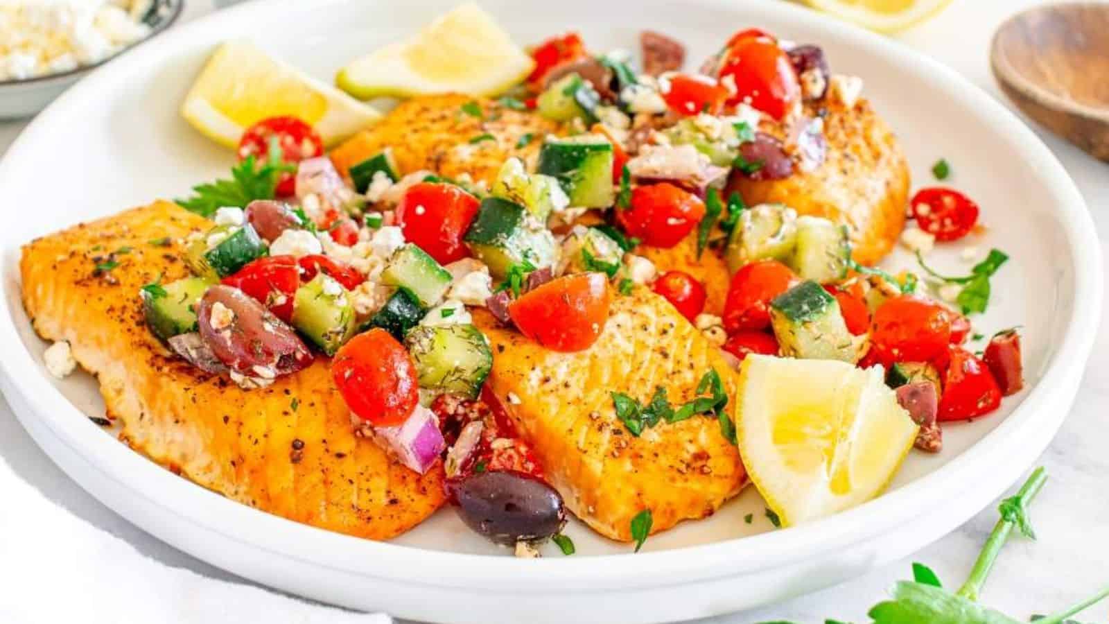 Grilled salmon fillets topped with a fresh cucumber, tomato, and olive salad, garnished with lemon wedges.
